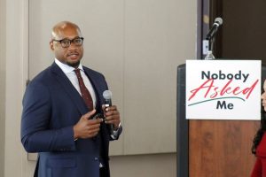 A person holding a microphone with a "Nobody Asked Me" sign on a podium behind them.